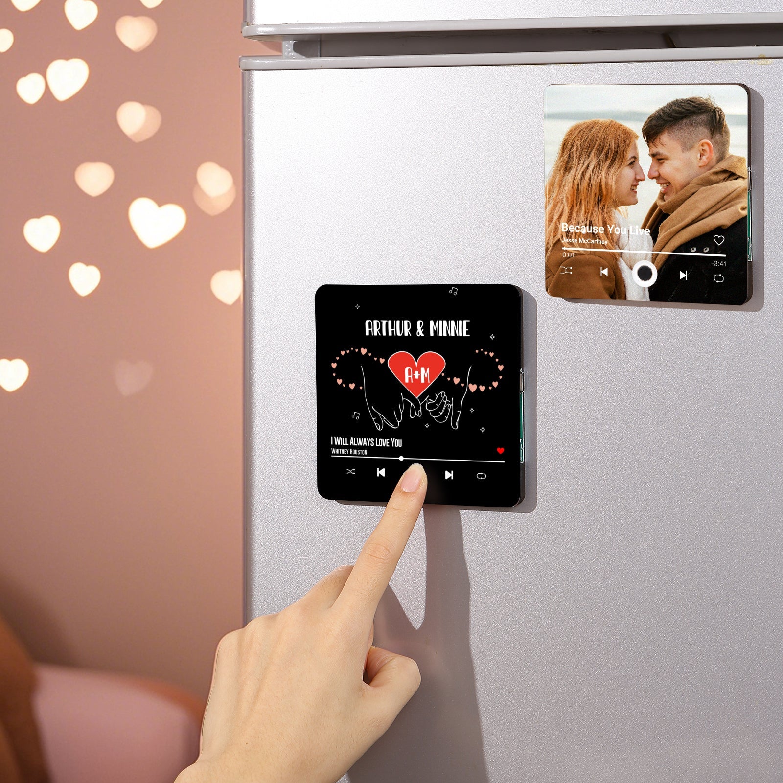 Close-up shot of personalized song plaque magnets on a refrigerator. One magnet depicts a couple in love, while the other features the names "Arthur & Minnie" with a heart symbol. A hand is shown interacting with the "I Will Always Love You" magnet.
